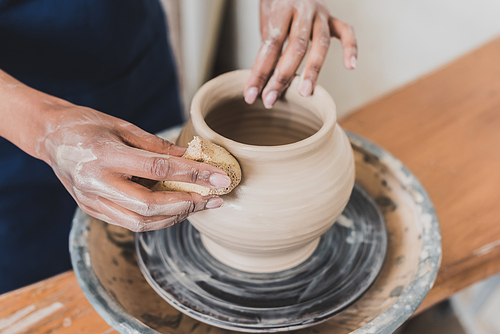 partial view of young african american woman modeling wet clay pot on wheel with hands and sponge in pottery