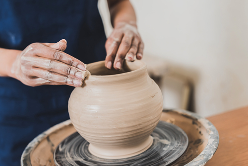 partial view of young african american woman modeling wet clay pot on wheel with hands in pottery