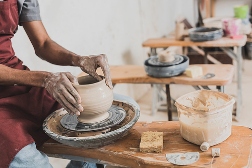 partial view of young african american man modeling wet clay pot on wheel with hands in pottery