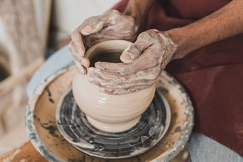 close up view of young african american man modeling wet clay pot on wheel with hands in pottery