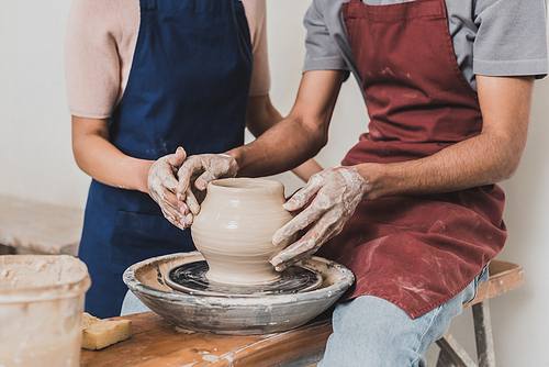 partial view of young african american couple modeling wet clay pot on wheel with hands in pottery