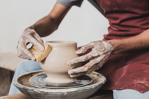 partial view of young african american man modeling wet clay pot on wheel with hands in pottery
