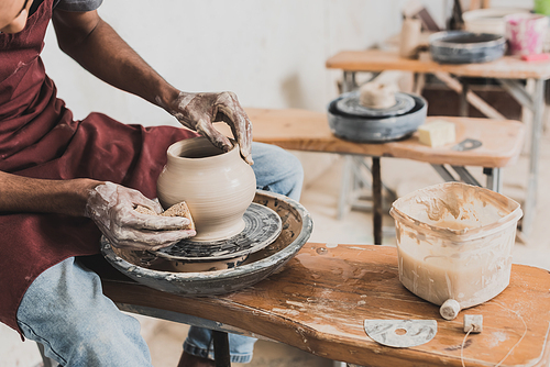 partial view of young african american man in casual clothes and apron modeling wet clay pot on wheel in pottery
