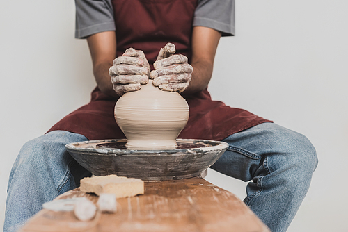 partial view of young african american man sitting on bench and shaping wet clay pot on wheel with hands in pottery