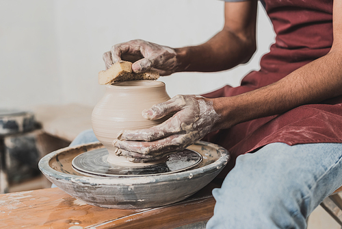 partial view of young african american man in jeans sitting on bench and shaping wet clay pot on wheel with sponge in pottery