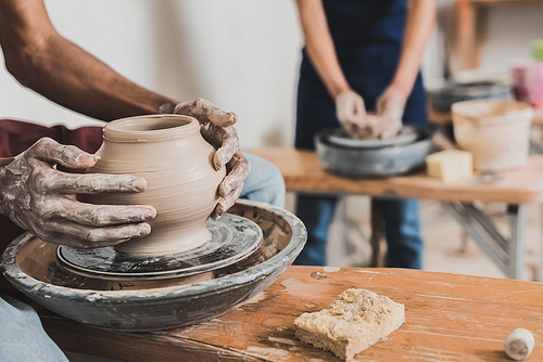 partial view of young african american man shaping wet clay pot on wheel near blurred woman in pottery