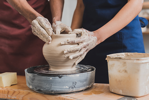 partial view of young african american couple shaping wet clay pot on wheel with hands in pottery