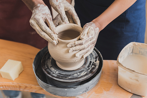 partial view of young african american couple shaping wet clay pot on wheel with hands in pottery