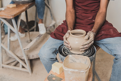 partial view of young african american man making wet clay pot on wheel in pottery