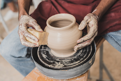 partial view of young african american man holding sponge and making wet clay pot on wheel in pottery