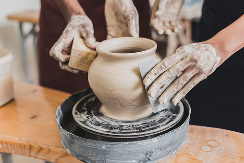 partial view of young african american couple shaping wet clay pot on wheel with hands and scraper in pottery
