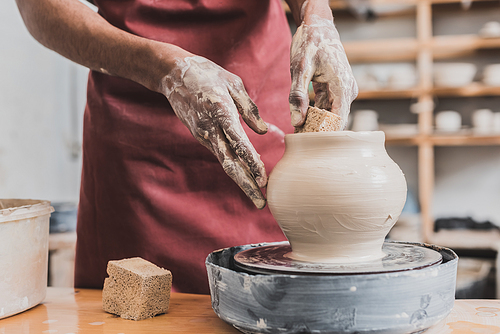 partial view of young african american man shaping wet clay pot with sponge on wheel in pottery