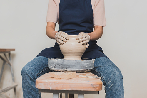 partial view of young african american woman in apron sitting on bench and shaping wet clay pot in pottery