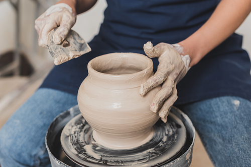 partial view of young african american woman in apron sitting on bench and shaping wet clay pot with scraper in pottery