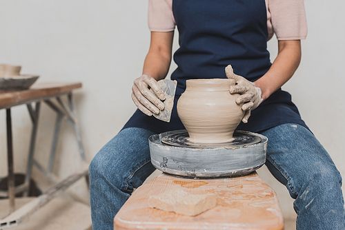 partial view of young african american woman sitting on bench and shaping wet clay pot with scraper in pottery