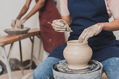 partial view of young african american woman shaping wet clay pot on wheel with scraper in pottery