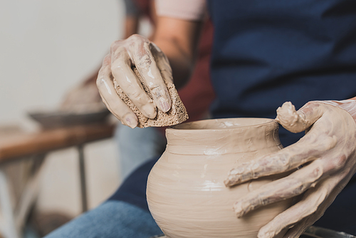partial view of female african american hands shaping wet clay pot on wheel with sponge in pottery
