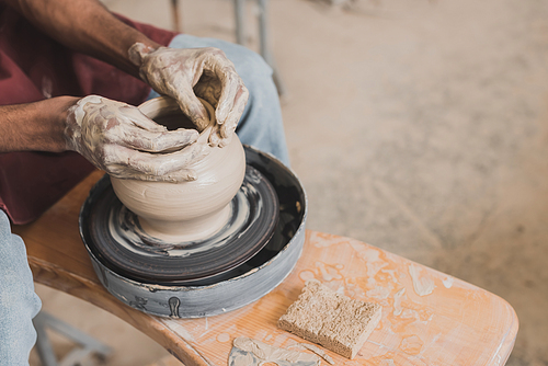 partial view of male african american hands shaping wet clay pot on wheel near sponge and scraper in pottery