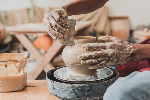 close up view of young african american man in apron sculpting pot on wheel with scraper in hand in pottery