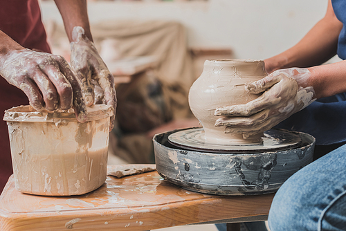 partial view of young african american couple sculpting pot on wheel near plastic box with water in pottery