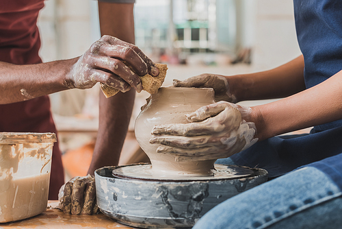 partial view of young african american couple in aprons sculpting pot on wheel with wet sponge in pottery