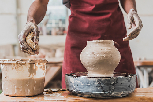 partial view of young african american man in apron sculpting clay pot on wheel and squeezing wet sponge in pottery