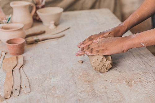 partial view of female african american hands rolling out piece of clay on wooden table in pottery