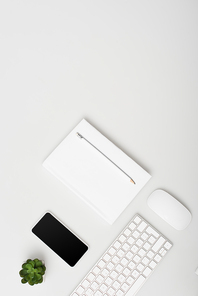 top view of smartphone with blank screen near small plant, keyboard and notebook with pencil isolated on white