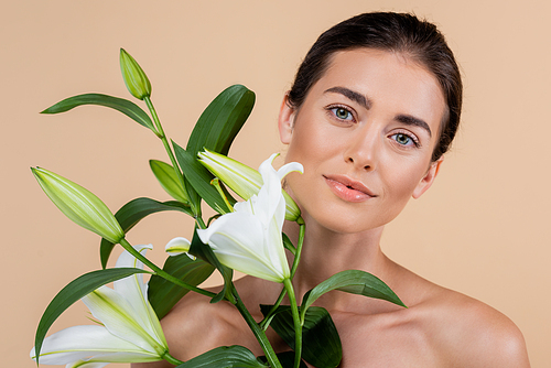 young woman with clean skin and naked shoulders near lily flowers isolated on beige, beauty concept