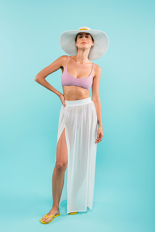 young woman in swimsuit, white pareo and sun hat posing with hand on hip on blue, beauty concept