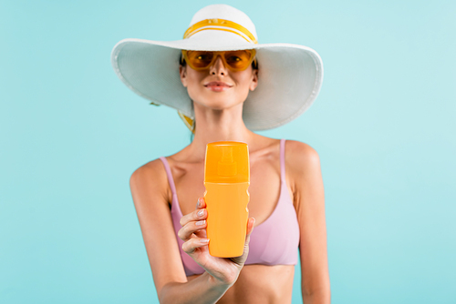 blurred woman in sun hat and orange sunglasses showing sunblock bottle isolated on blue
