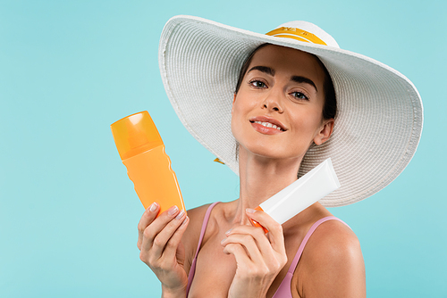 joyful woman in sun hat posing with tube and bottle of sunblock isolated on blue