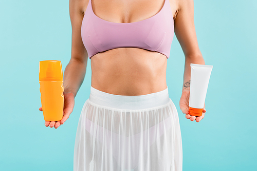 cropped view of woman in swimsuit and pareo holding tube and bottle of sunscreen isolated on blue