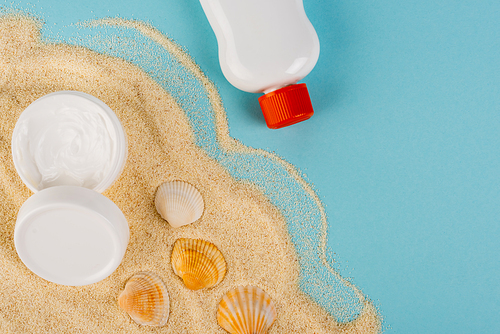 top view of sunscreen and cosmetic cream near seashells on sand and blue surface