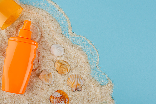 top view of orange sunblock bottle near seashells and sand on blue surface