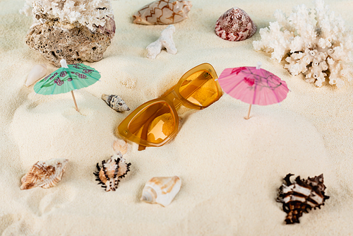 top view of orange sunglasses near seashells and cocktail umbrellas on sand