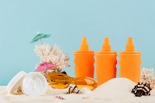 containers with sunscreen and cosmetic cream near sunglasses on sand isolated on blue