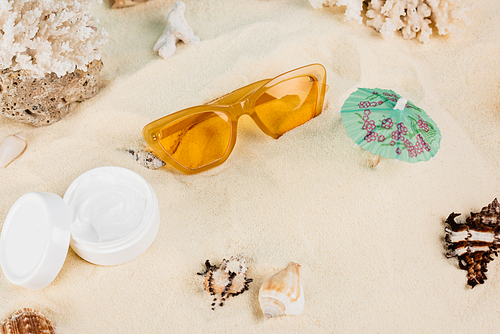 top view of cosmetic cream near orange sunglasses and seashells on sand