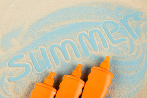 top view of orange bottles with sunblock near summer lettering on sand and blue surface