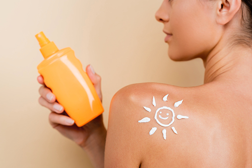 partial view of woman with image of happy sun on shoulder holding bottle of sunblock isolated on beige