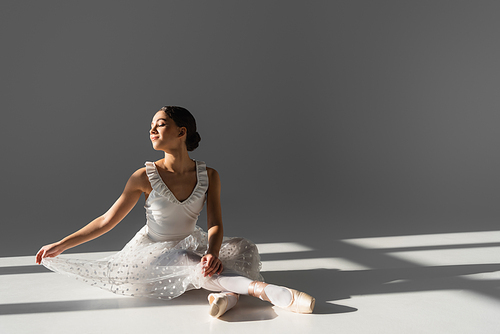 Side view of smiling elegant ballerina sitting on grey background