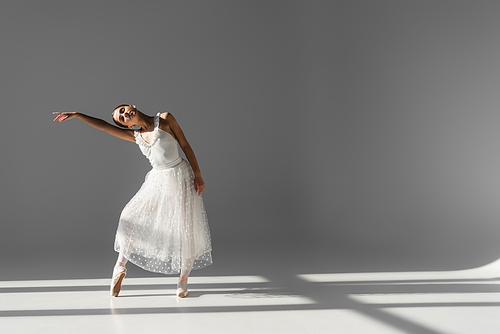 Ballerina dancing with closed eyes on grey background with sunlight