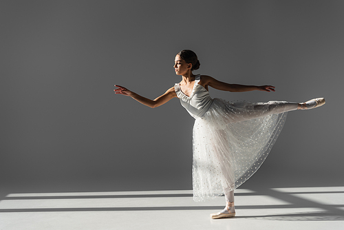 Side view of ballerina in white skirt dancing on grey background with sunlight