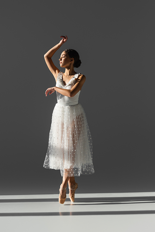 Side view of young ballerina with closed eyes dancing on grey background with sunlight