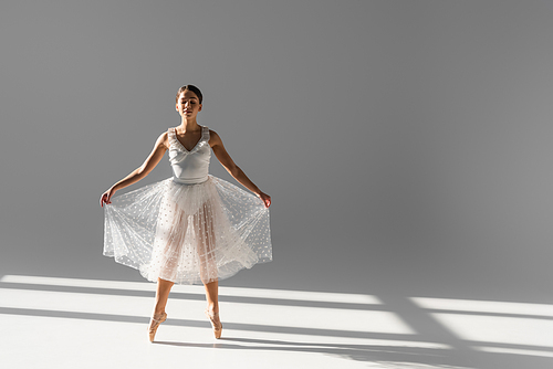 Full length of ballerina in skirt and pointe shoes dancing on grey background with sunlight