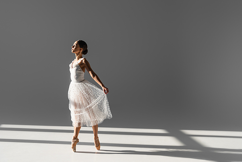 Side view of ballerina holding skirt while dancing on grey background with sunlight