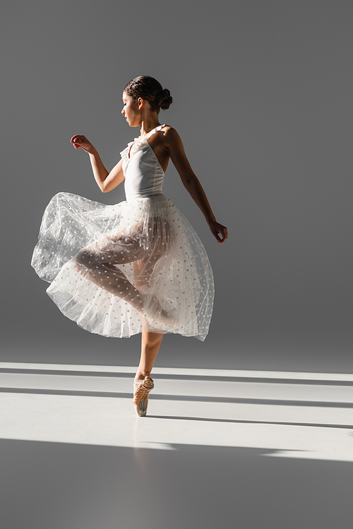 Side view of elegant ballerina in white skirt dancing on grey background with sunlight