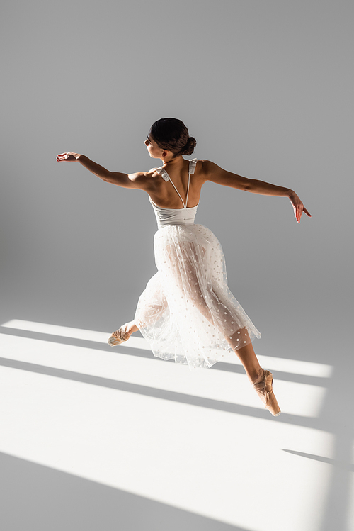 Side view of elegant ballerina jumping in sunlight on grey background