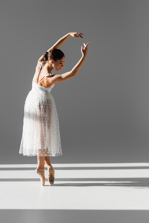 Side view of ballerina bending and looking down on grey background with sunlight