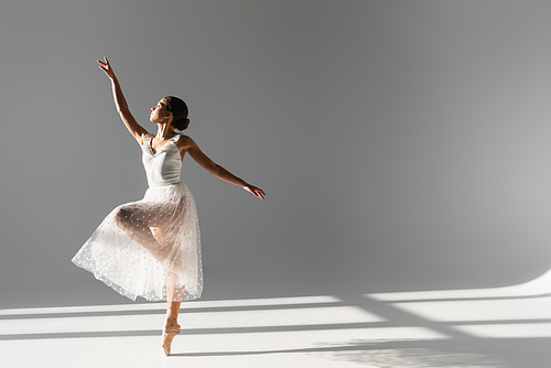 Side view of ballerina in white skirt standing on one leg on grey background with sunlight